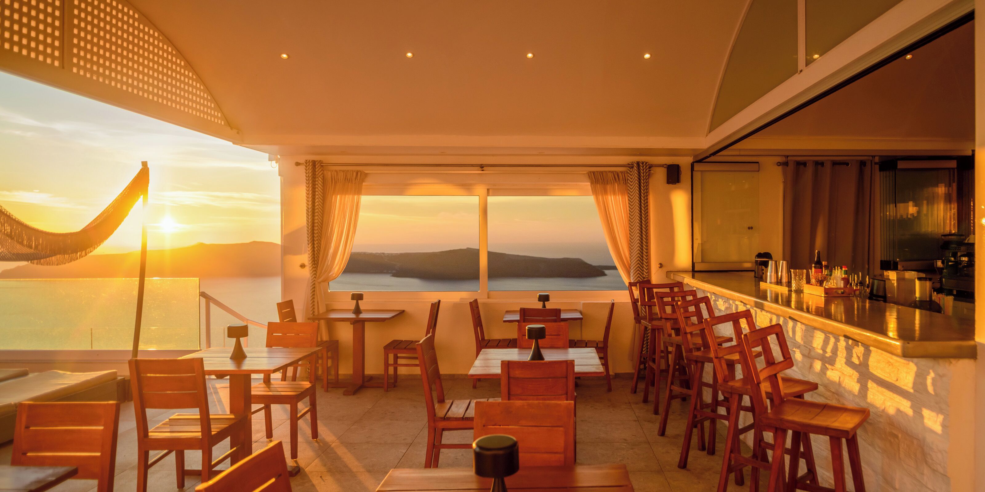 A restaurant with a view of the sunset over the water, wooden tables and chairs, and a bar area.