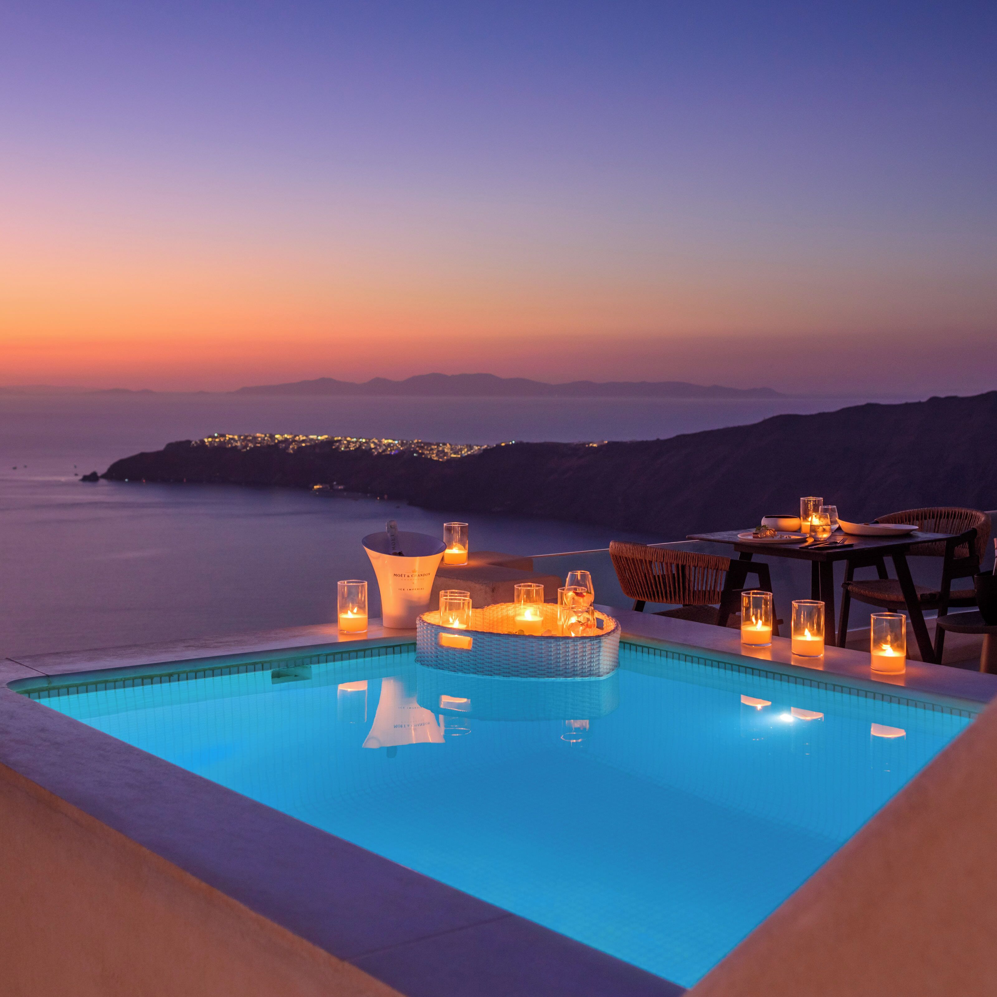 Una piscina con vistas al atardecer, una zona de comedor con sillas y una mesa, y un centro de mesa iluminado con velas.