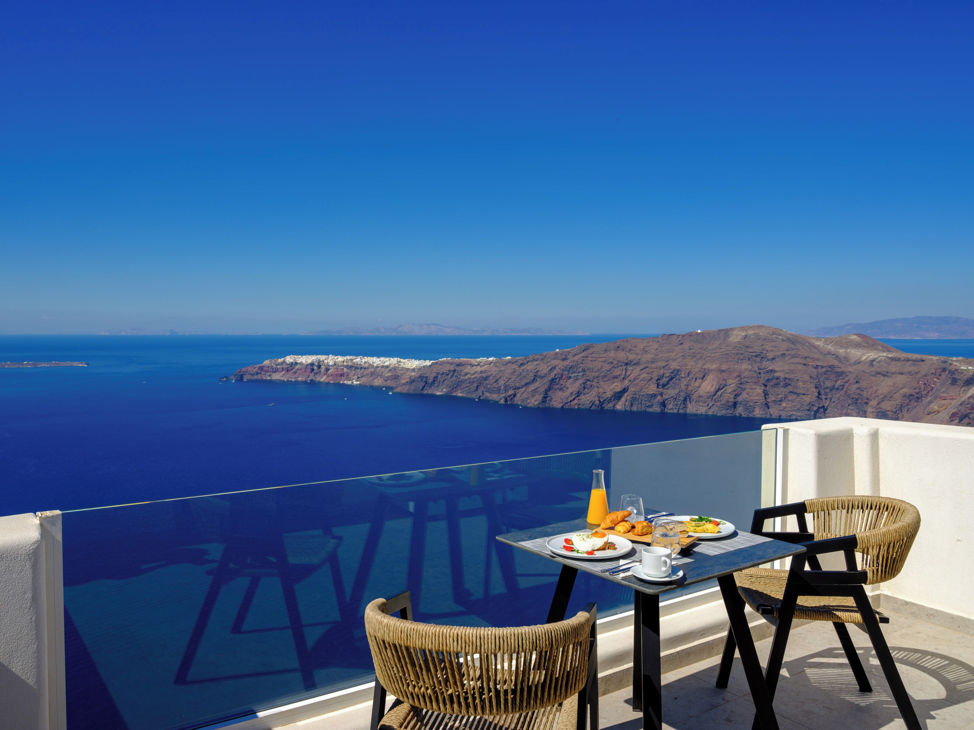 Una mesa con alimentos para el desayuno y sillas en un balcón con vistas al mar y a los acantilados.