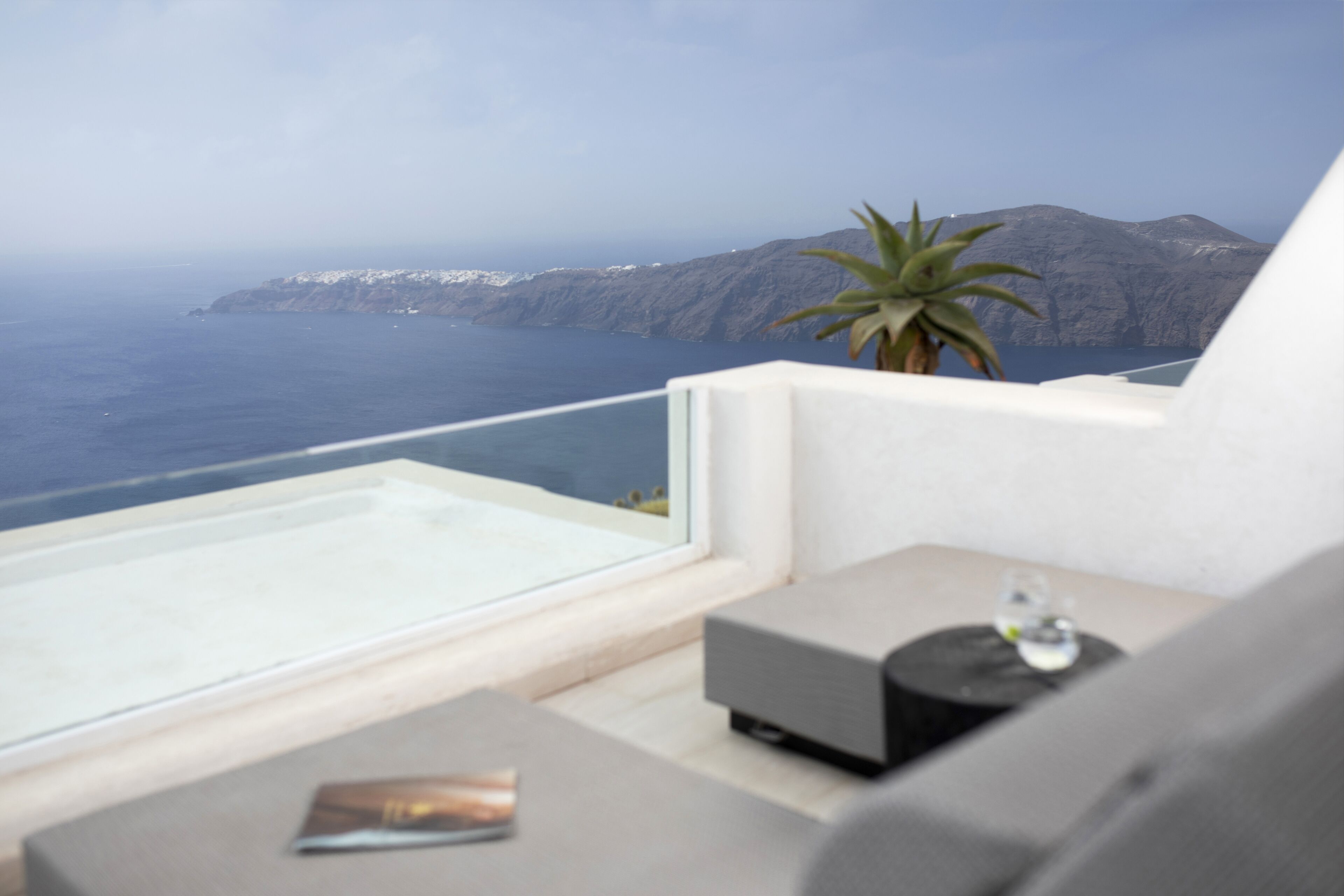 A balcony with a view of the sea and mountains, a plant, and a small table with a decorative item.