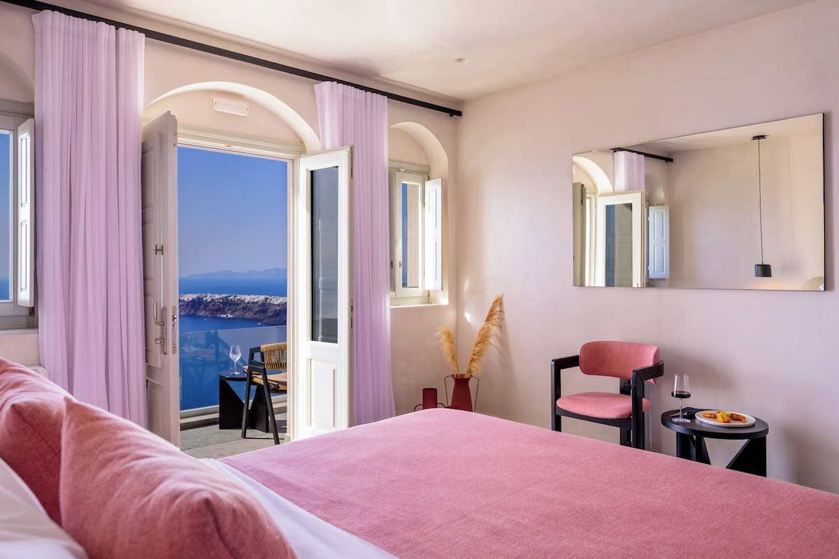 A hotel room with a large bed, a chair, a small table with a plate of food and a glass of wine, and a view of the sea through an open door.