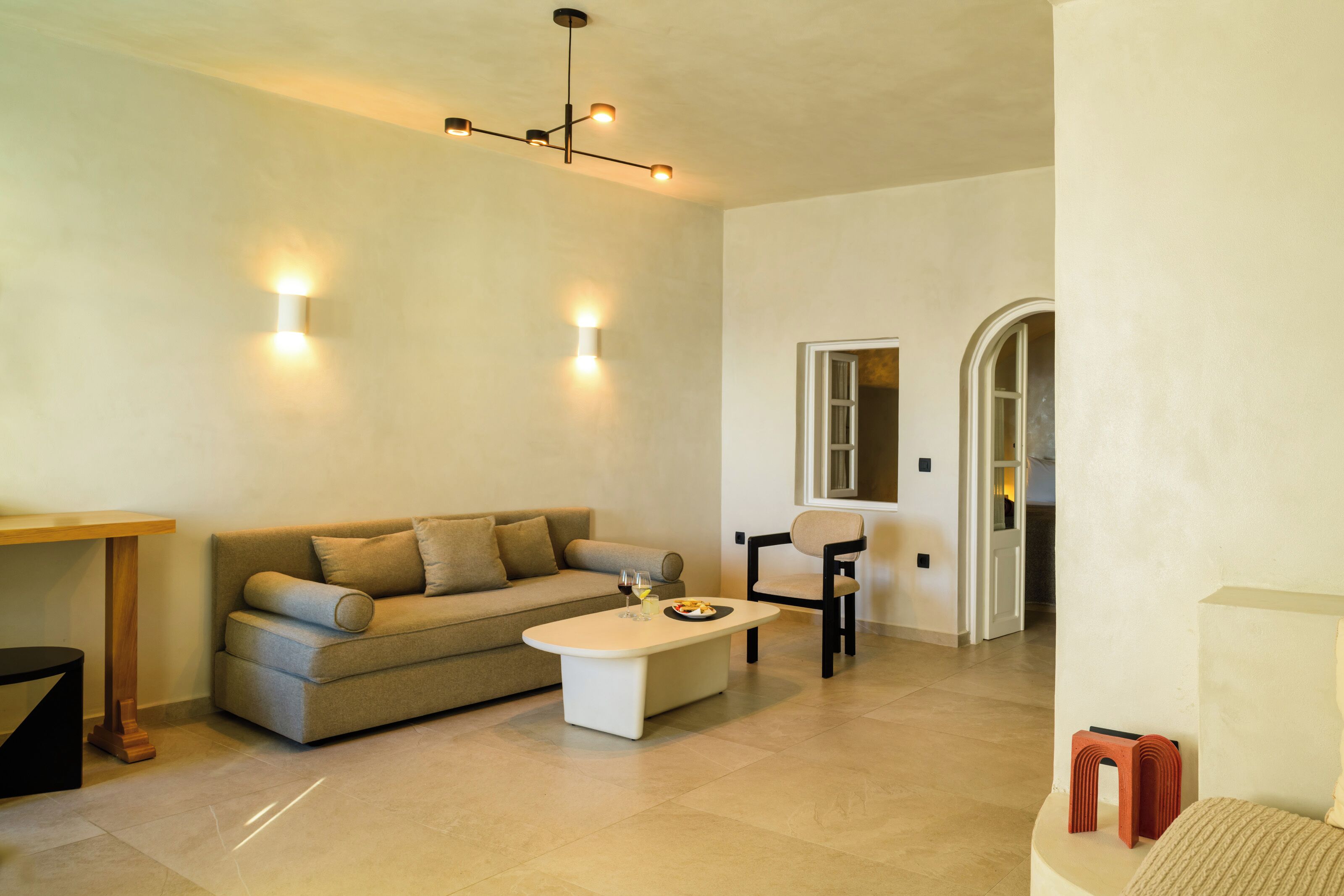 Una sala de estar moderna con un sofá, una mesa de centro y una silla. Cuenta con una mesa de comedor y un taburete pequeño. El piso y las paredes son de color claro, y hay una lámpara en el techo.