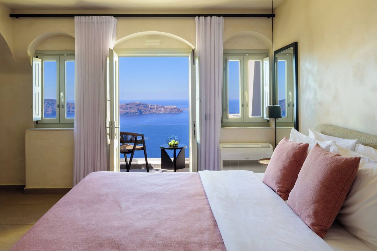 A hotel room with a bed, two pillows, a chair, a small table with a fruit bowl, and a view of the sea through open windows.