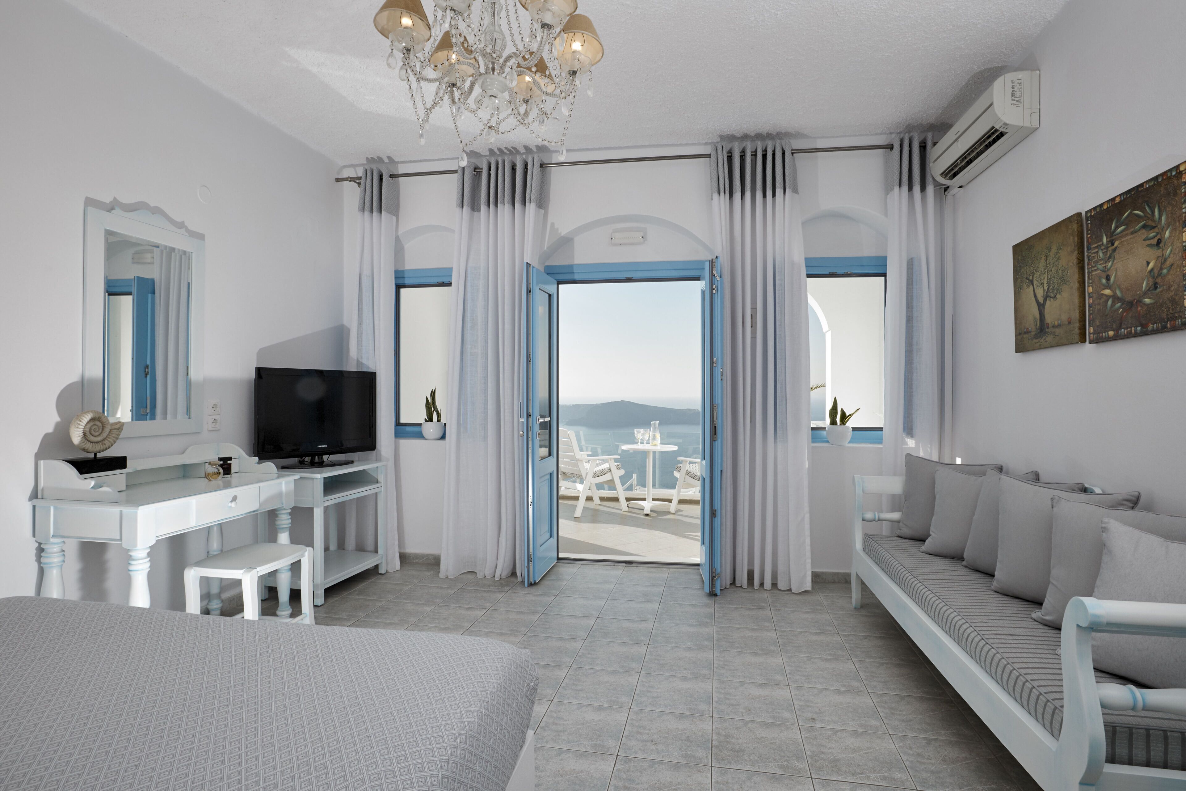 A modern hotel room with a large bed, a desk with a television, a seating area with cushions, and a view of the sea through open doors.