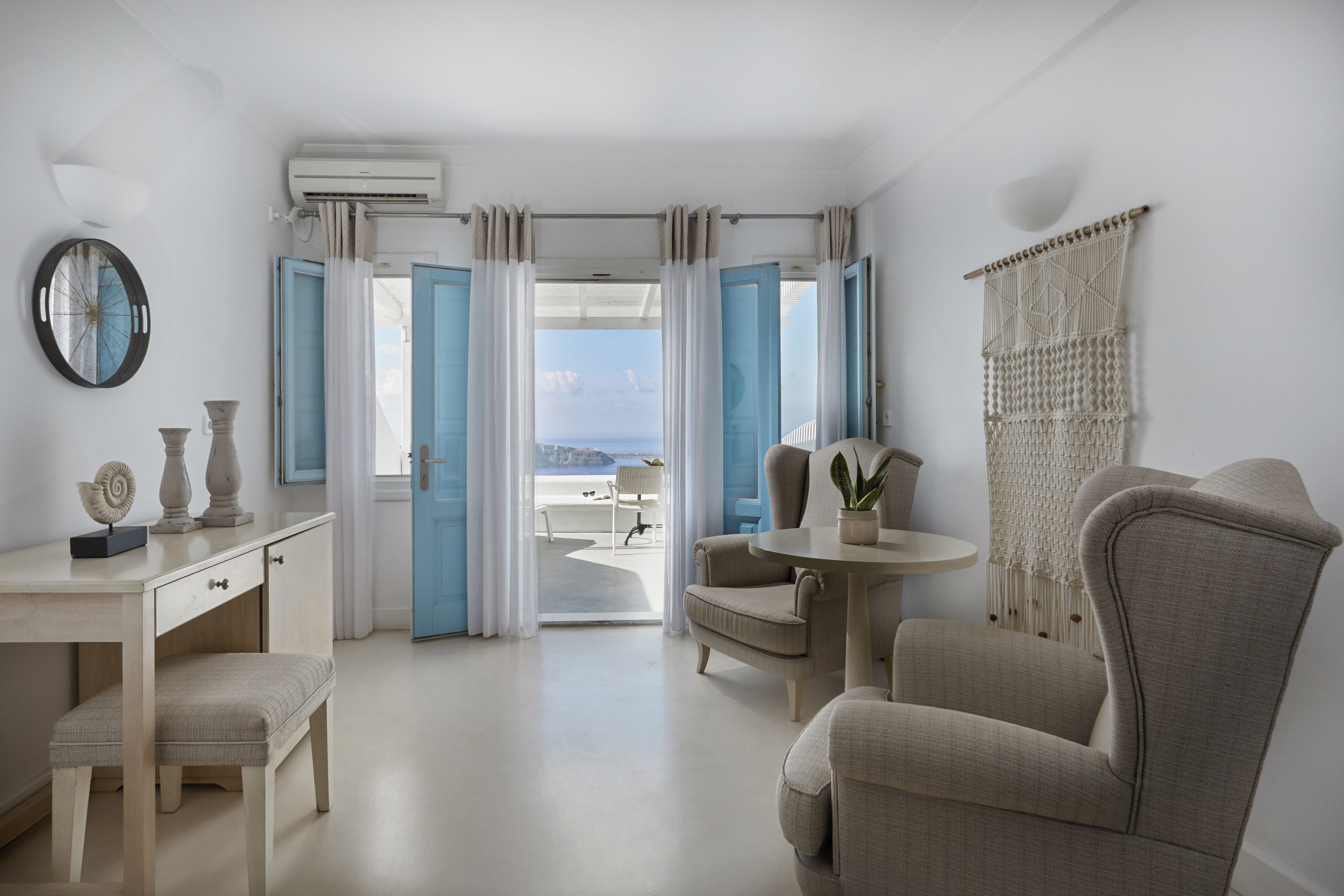 A room with a view of the sea, a desk with a chair, a round table, and a macrame wall hanging.
