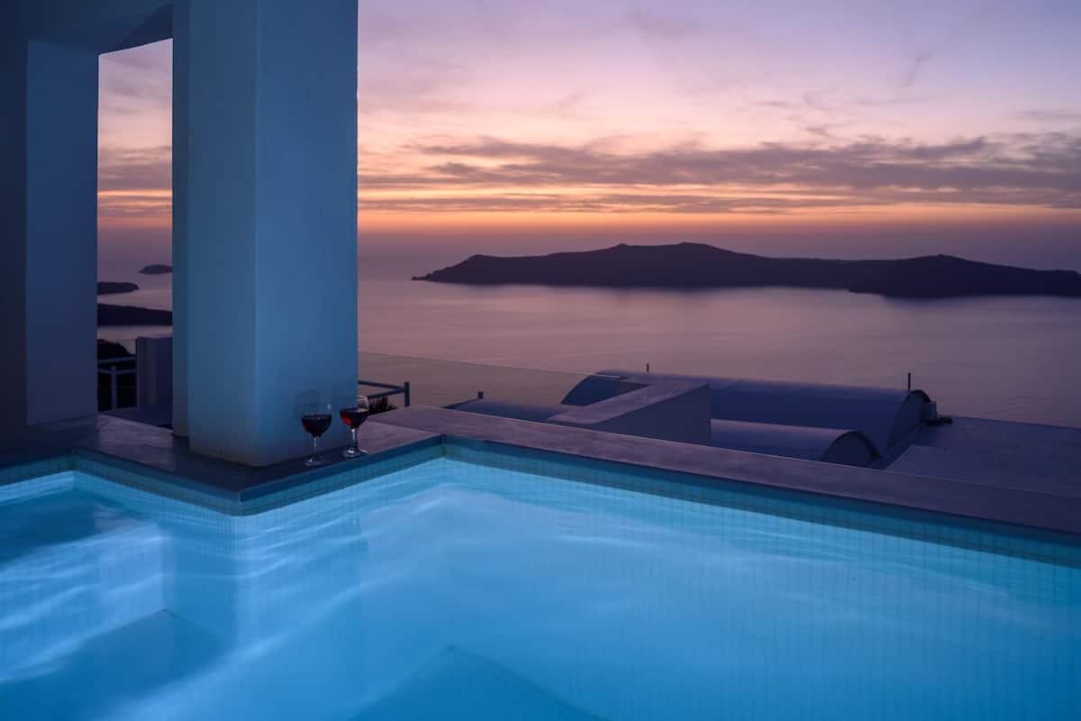 A pool with a view of a sunset over water, a pillar, and two glasses of red wine.