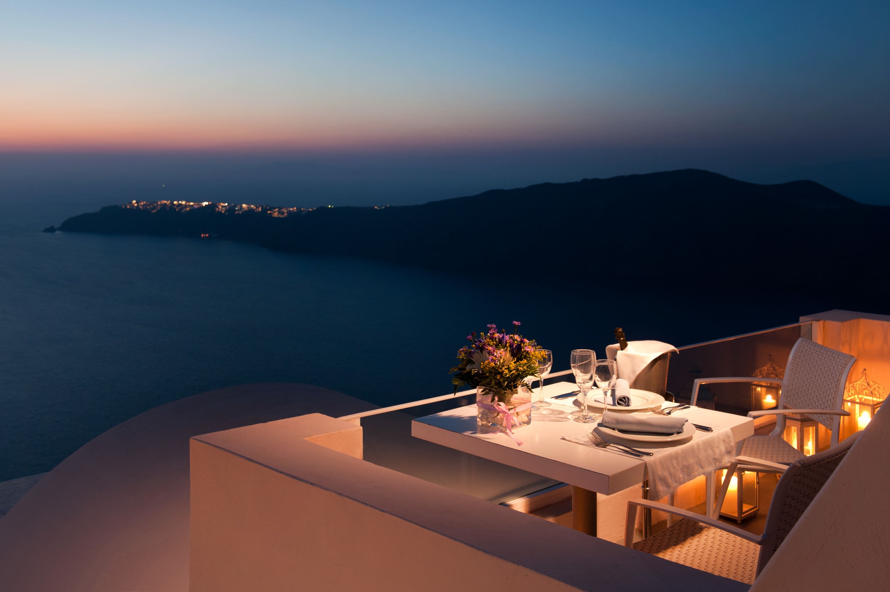 Une table dressée pour deux personnes, ornée d’un vase de fleurs, d’un verre à vin et d’une bougie, sur un balcon donnant sur la mer au crépuscule.