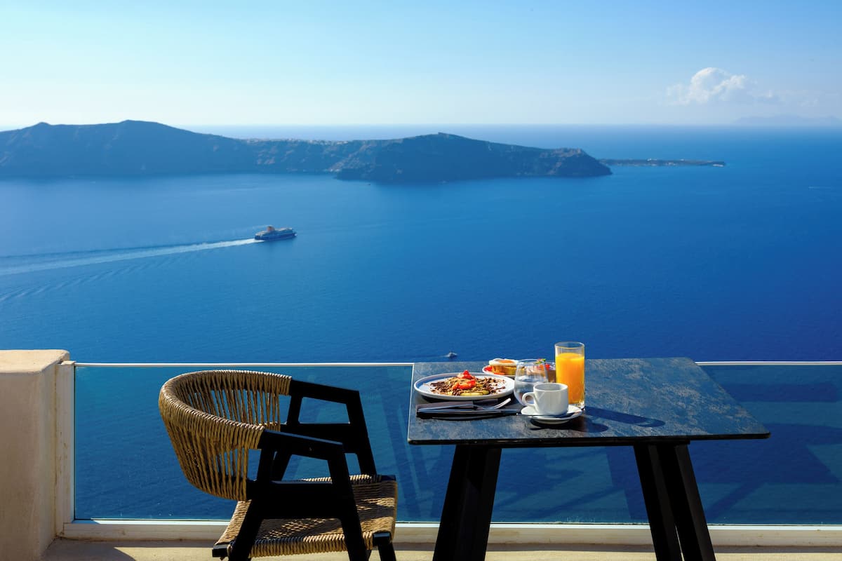 A table with breakfast and a wicker chair, overlooking a sea with a ship in the distance and a mountainous coastline.