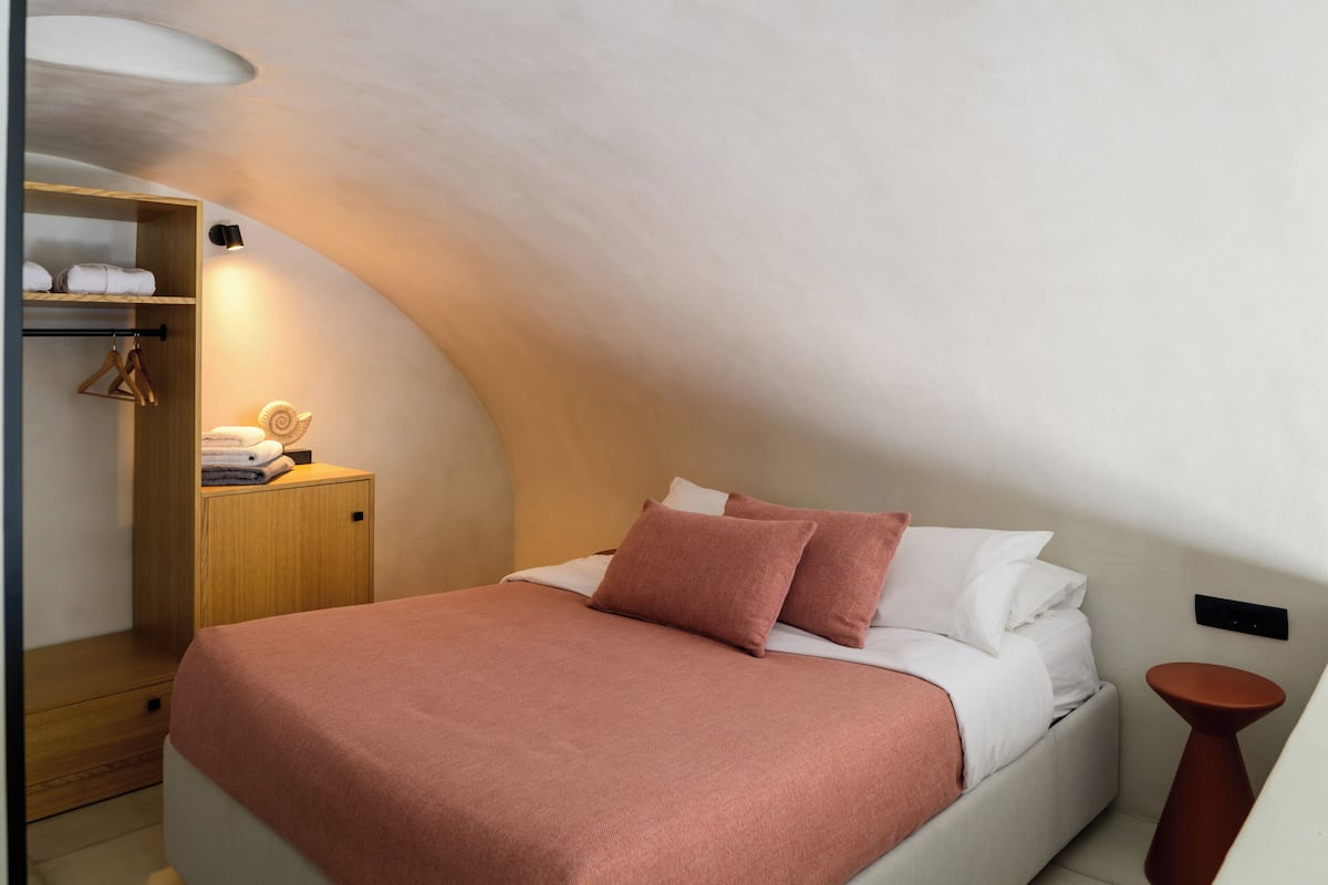 A bedroom with a curved ceiling, a bed with a pink blanket, white pillows, and a wooden nightstand with a lamp.