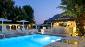 Piscina con sillas de descanso y sombrillas, rodeada de árboles y una zona de bar.