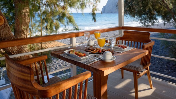 Una mesa de madera con alimentos para el desayuno y un vaso de jugo de naranja en un balcón con vista al mar.
