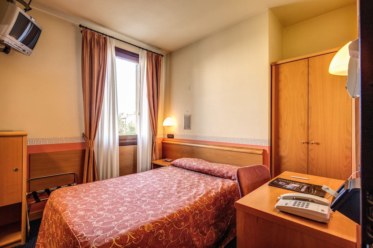 A hotel room with a bed, a desk with a telephone, a wardrobe, and a television on the wall.