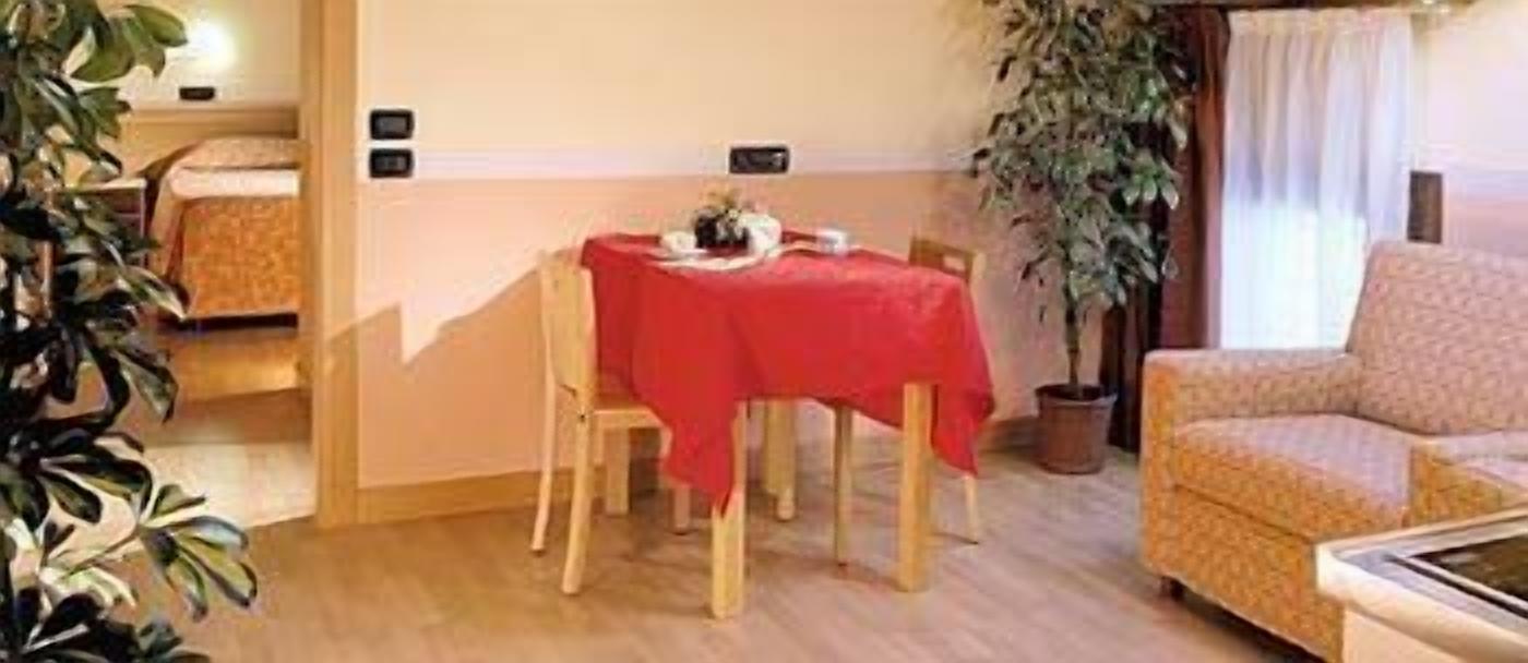 Ein gemütlicher Raum mit Esstisch, gedeckter Tischdecke rot, einer Couch und einem Topf mit Pflanze.
