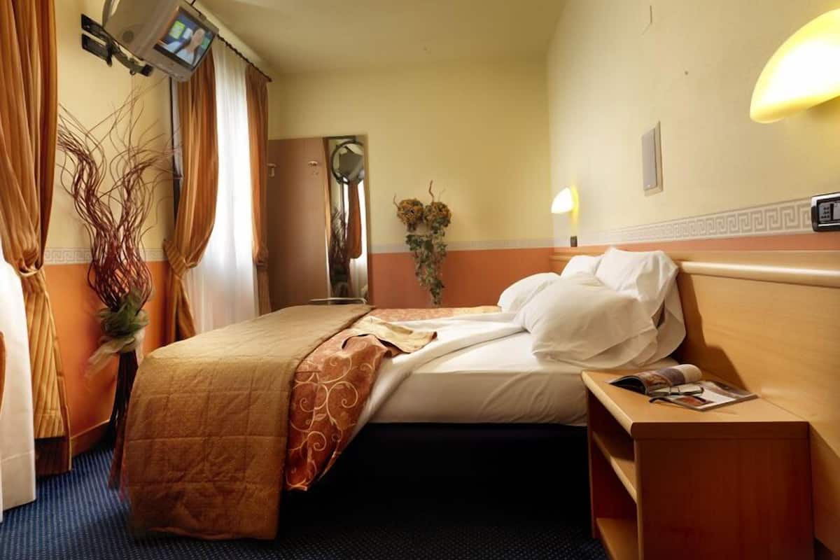 A hotel room with a bed, bedside table, lamp, curtains, and a television mounted on the wall.