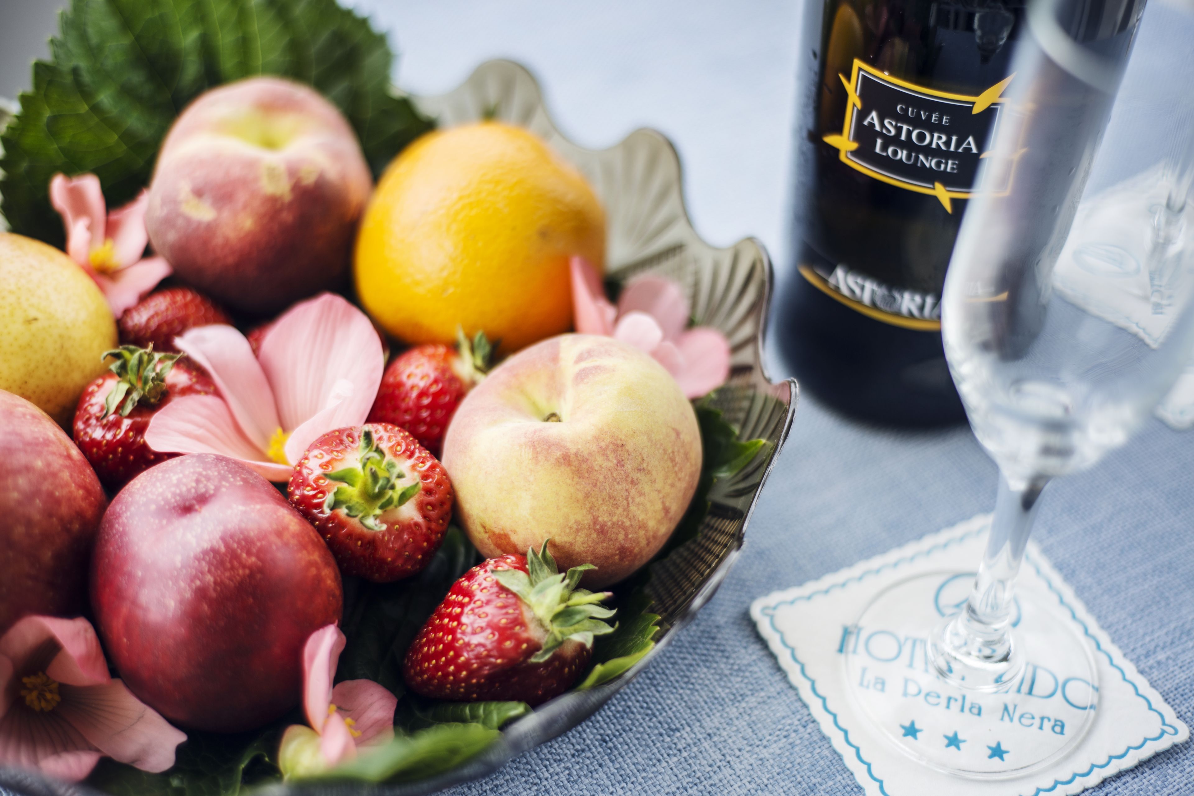 Un cuenco de fruta con fresas, melocotones y una naranja, junto a una botella de Astoria y una copa de vino.