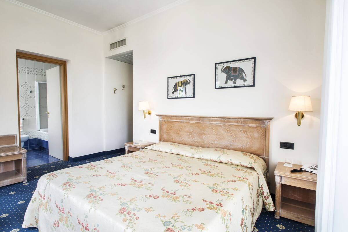 Een hotelkamer met een bed, twee wandlampen, een nachtkastje, een houten bankje en twee ingelijste foto's aan de muur.
