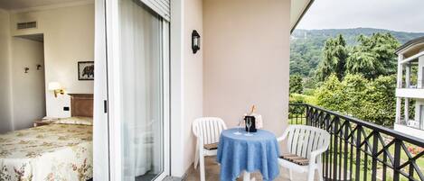 A balcony with a bed, a table with a bottle and glasses, and a view of greenery and hills.