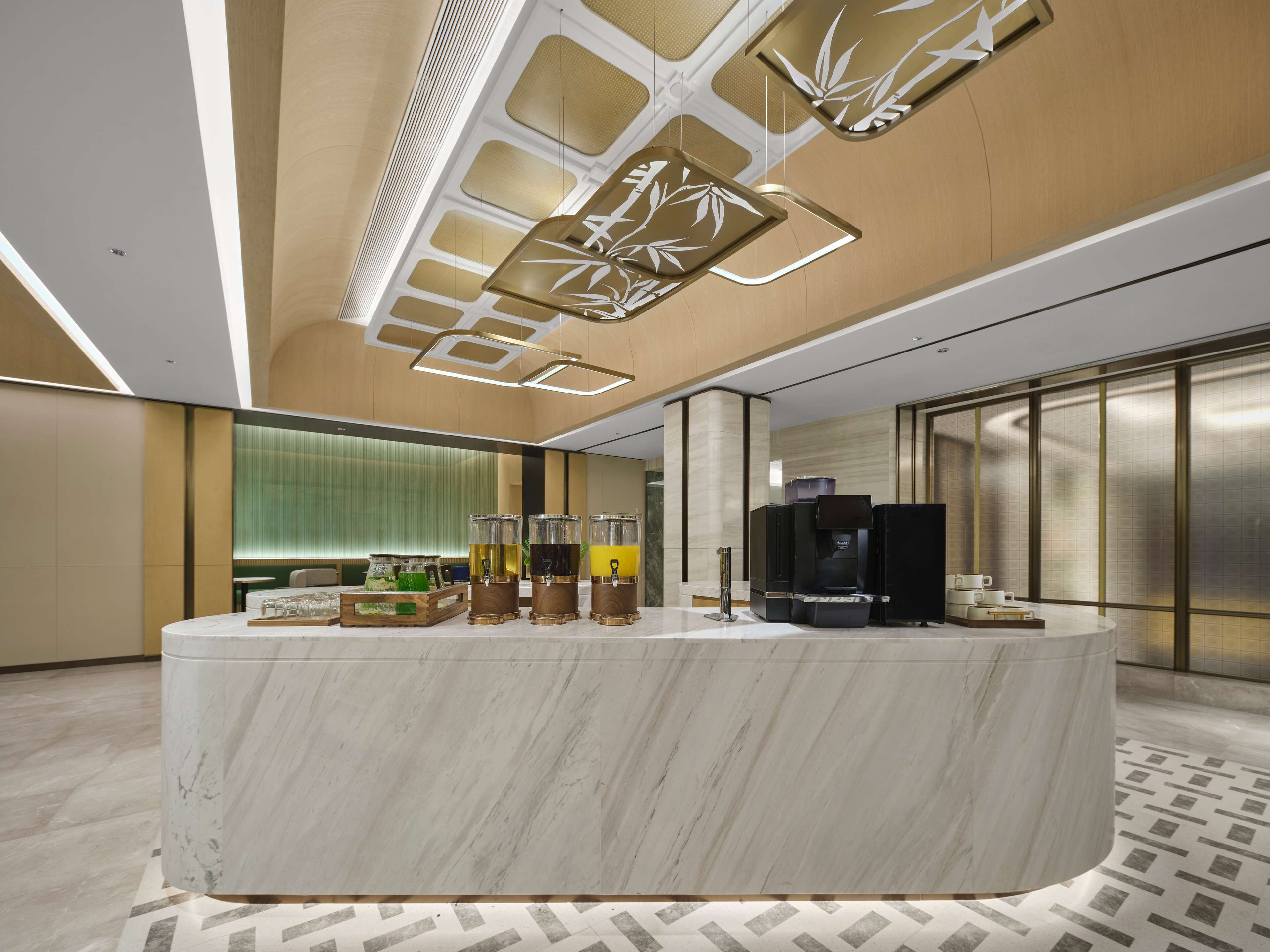 A modern hotel lobby with a marble reception desk, beverage dispensers, a television, and a patterned floor.