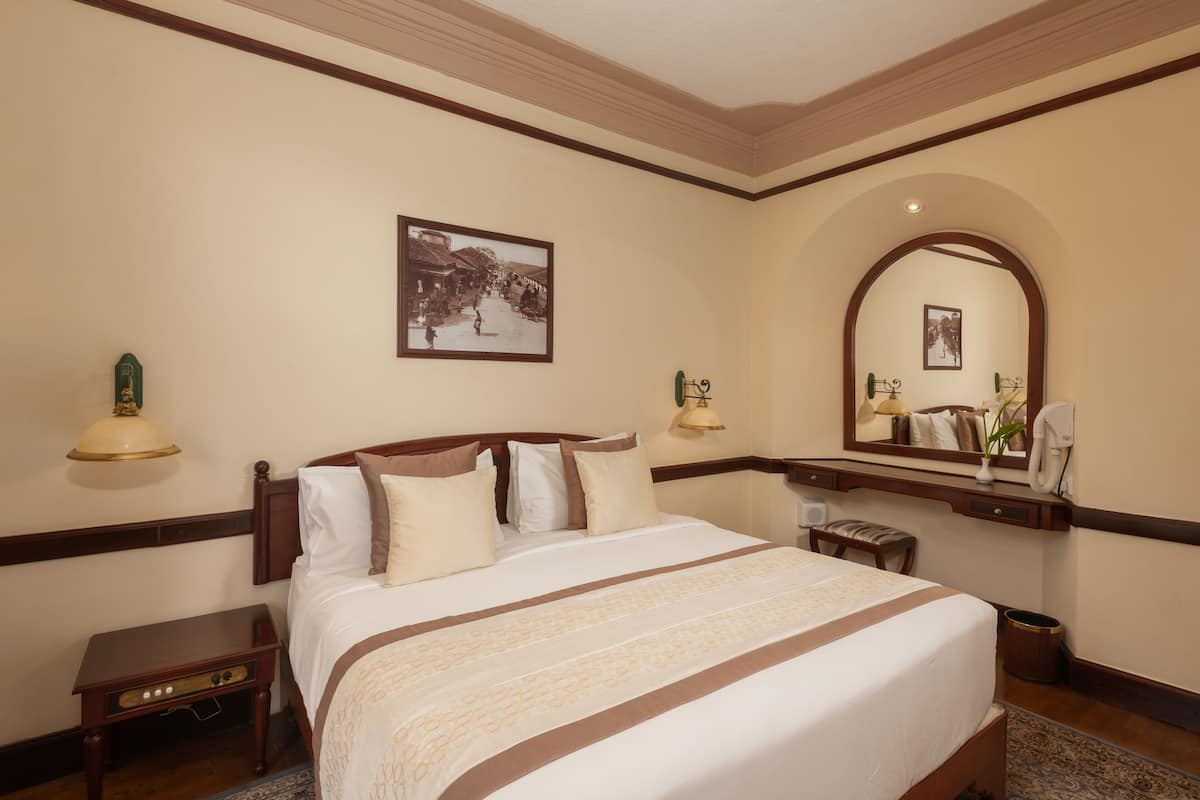 A hotel room with a large bed, a nightstand, a desk with a lamp, a mirror, and a picture on the wall.