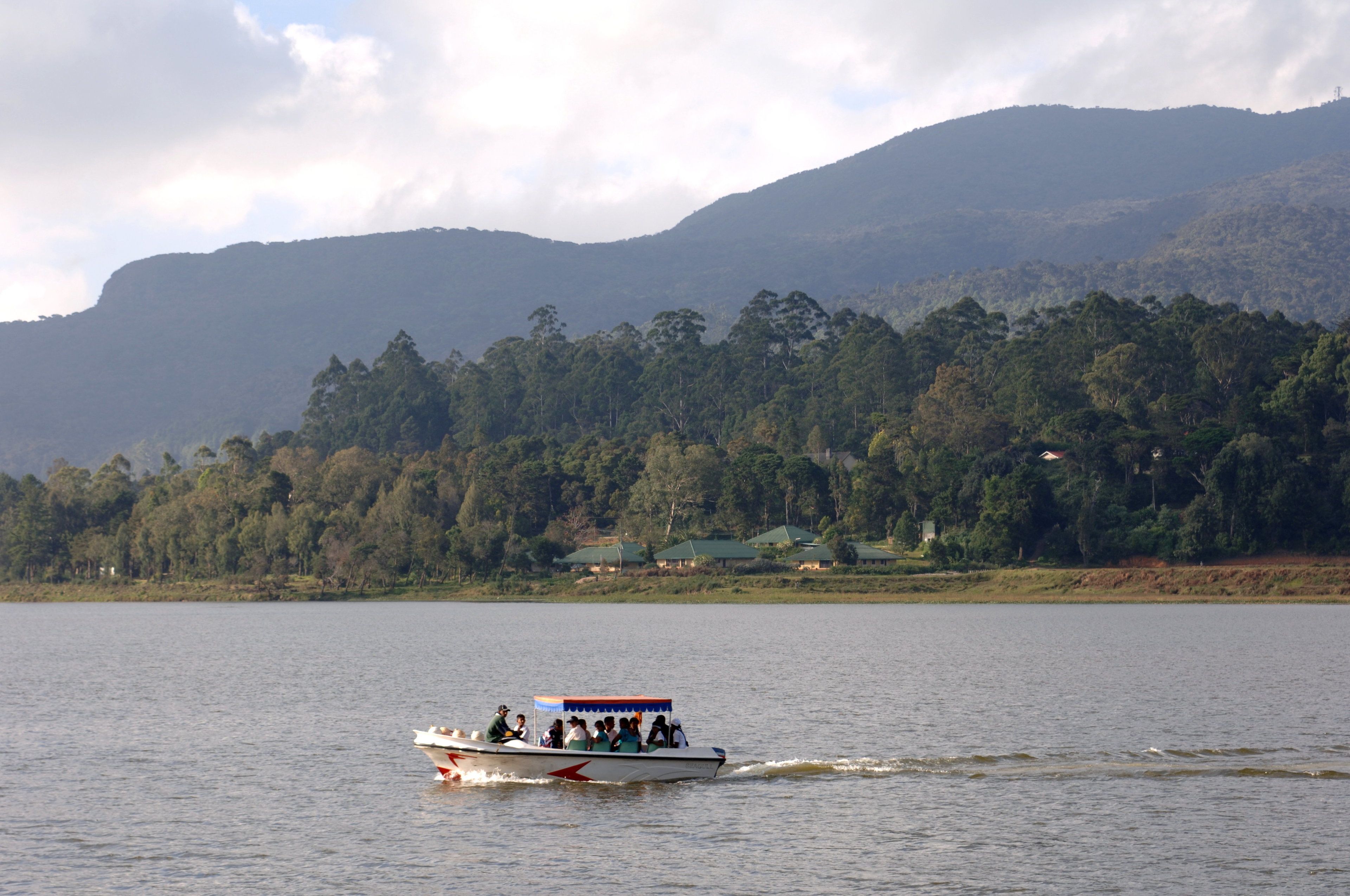 A boat with passengers on a calm lake, surrounded by dense forest and hills.