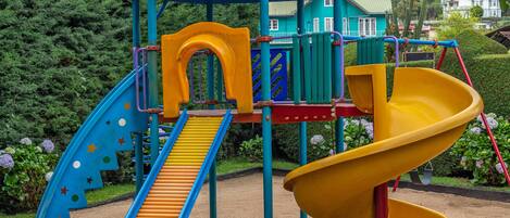 Un parque infantil con un tobogán azul y amarillo, rodeado de vegetación y colinas.