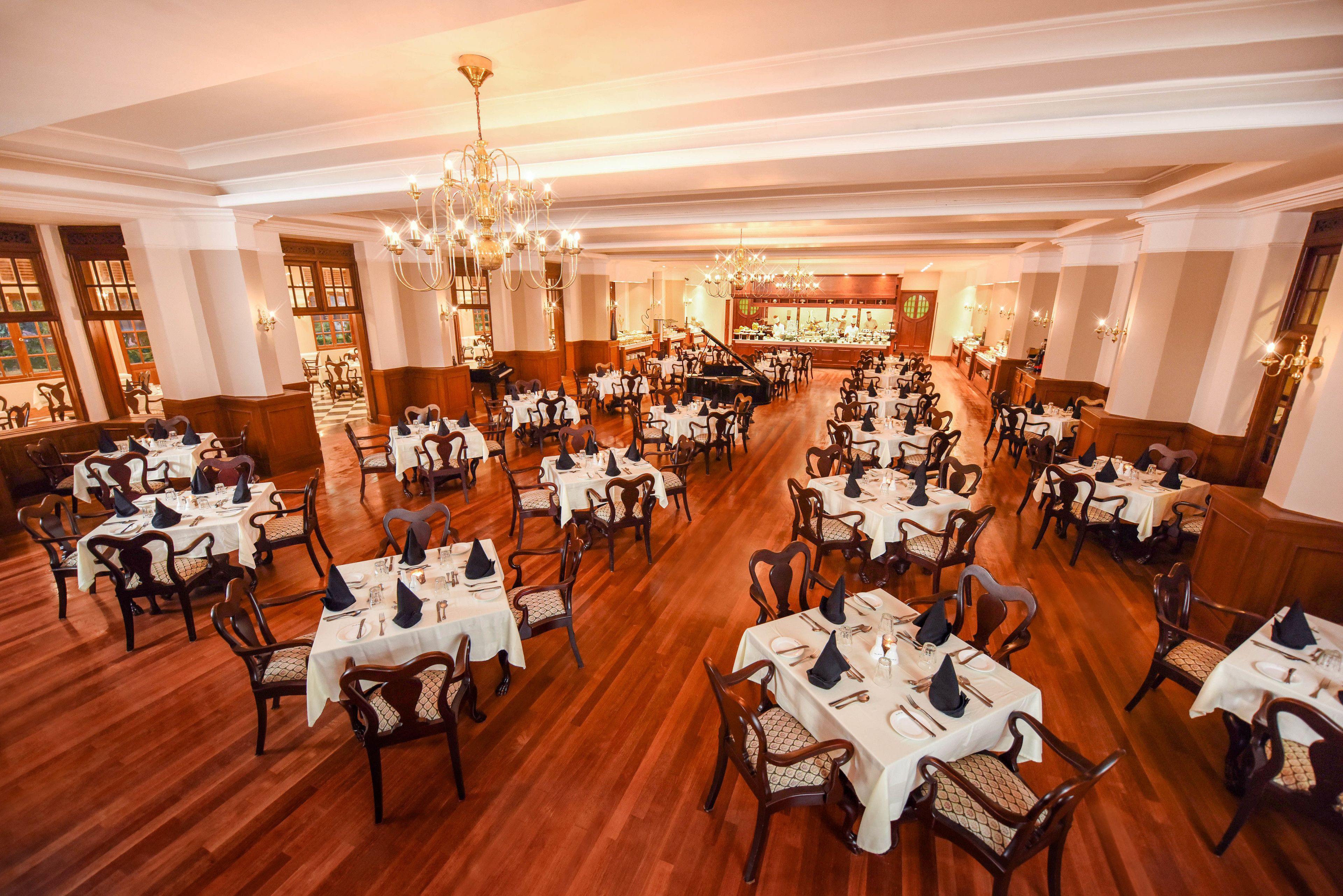 A spacious dining hall with multiple tables and chairs, chandeliers, and a piano.