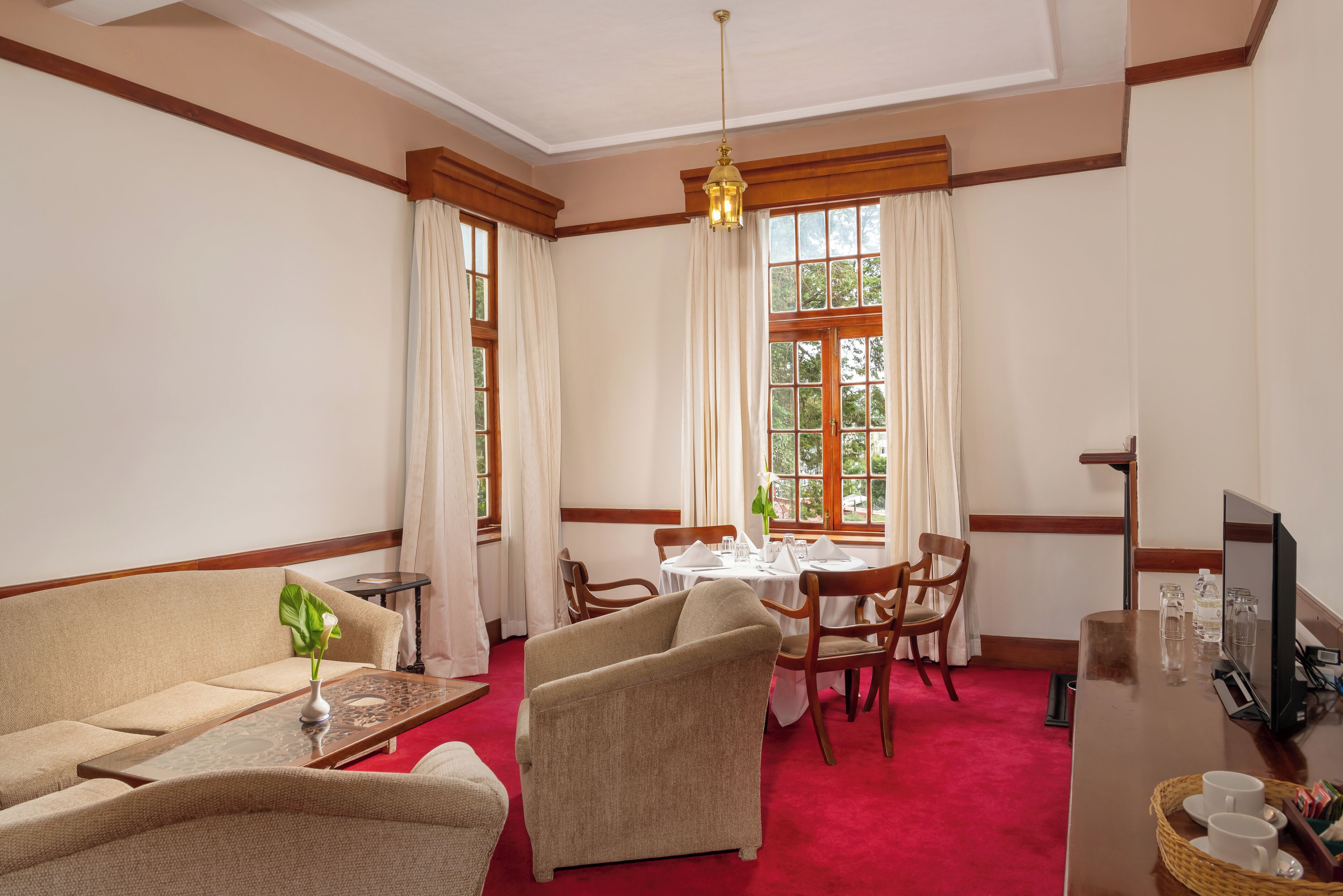 A hotel room with a red carpet, a sofa, two armchairs, a dining table with chairs, a TV, and a window with curtains.