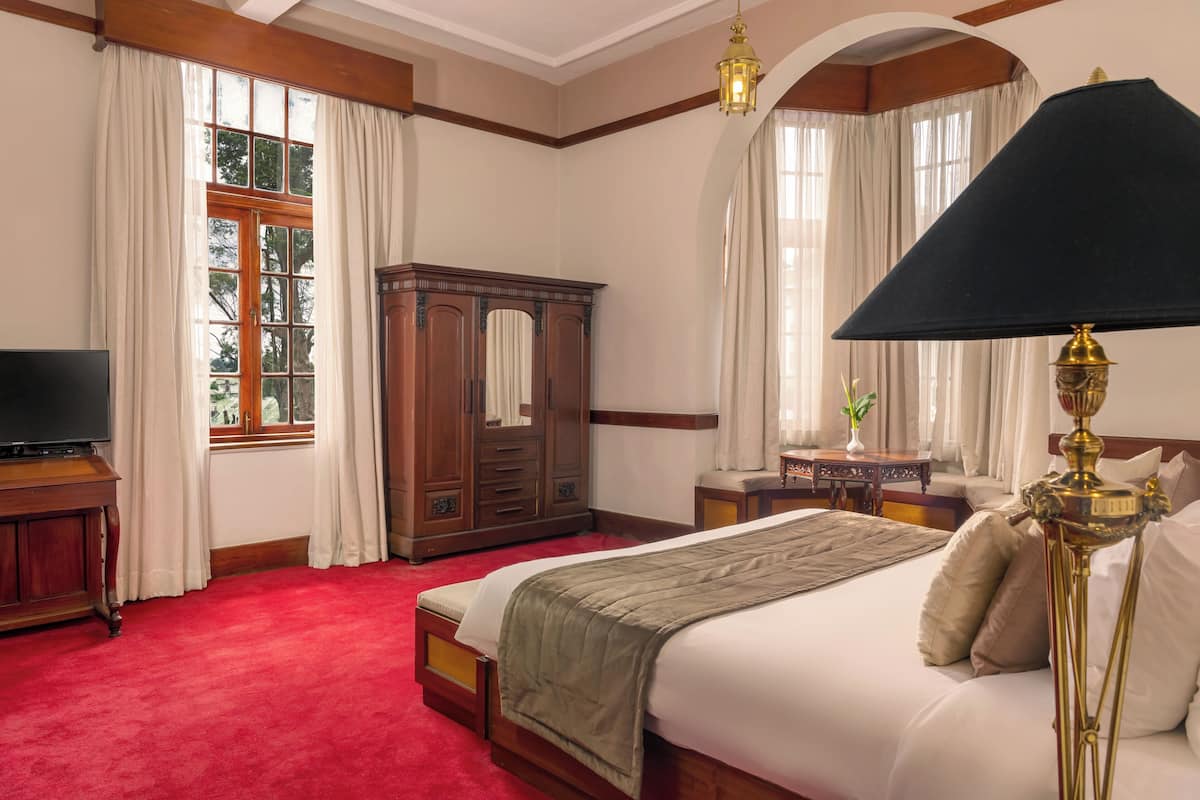 A hotel room with a large bed, a wooden wardrobe, a desk with a chair, a television, and a lamp.