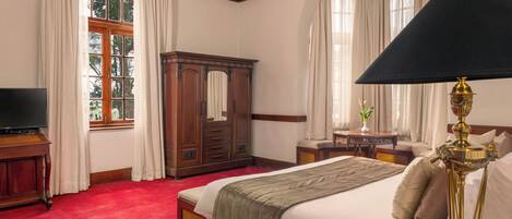 A hotel room with a large bed, a wooden wardrobe, a desk with a chair, a television, and a lamp.