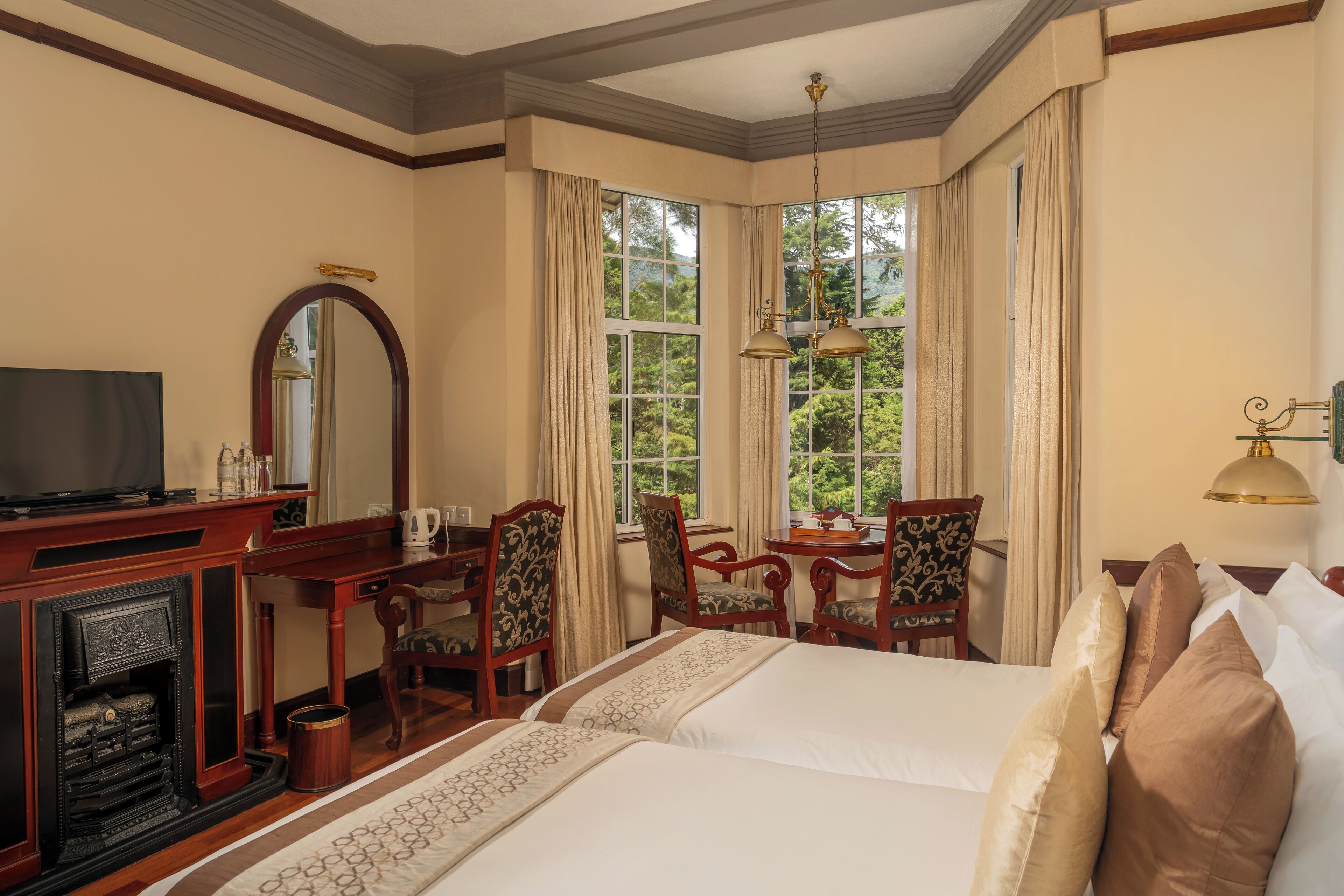 A hotel room with a large bed, a desk with a television, a fireplace, and a view of greenery through large windows.