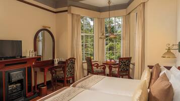 A hotel room with a large bed, a desk with a television, a fireplace, and a view of greenery through large windows.