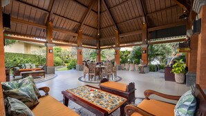 Un espace extérieur couvert, aménagé en patio, avec des meubles en bois, une table centrale recouverte d’une nappe à motifs floraux, et une vue sur la végétation environnante.