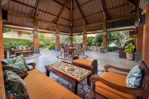 An outdoor covered patio area with wooden furniture, a central table with a floral tablecloth, and a view of the surrounding greenery.