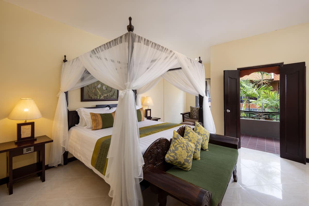 A four-poster bed with a canopy, two bedside tables with lamps, a green bedspread, and a view of a balcony with plants.