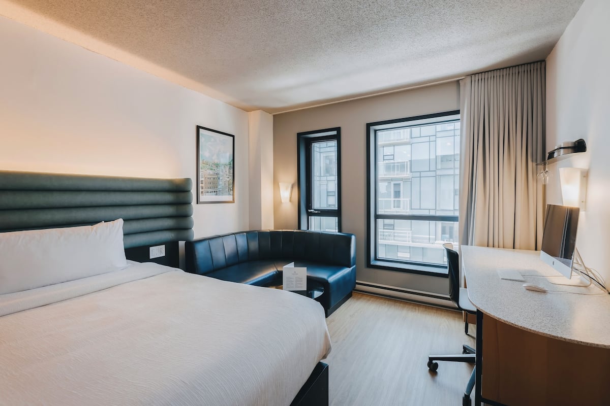 Une chambre d’hôtel avec un lit, un bureau, une chaise, un canapé et de grandes fenêtres.