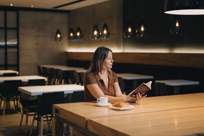 A woman reading a book at a wooden table in a modern cafe with pendant lighting.
