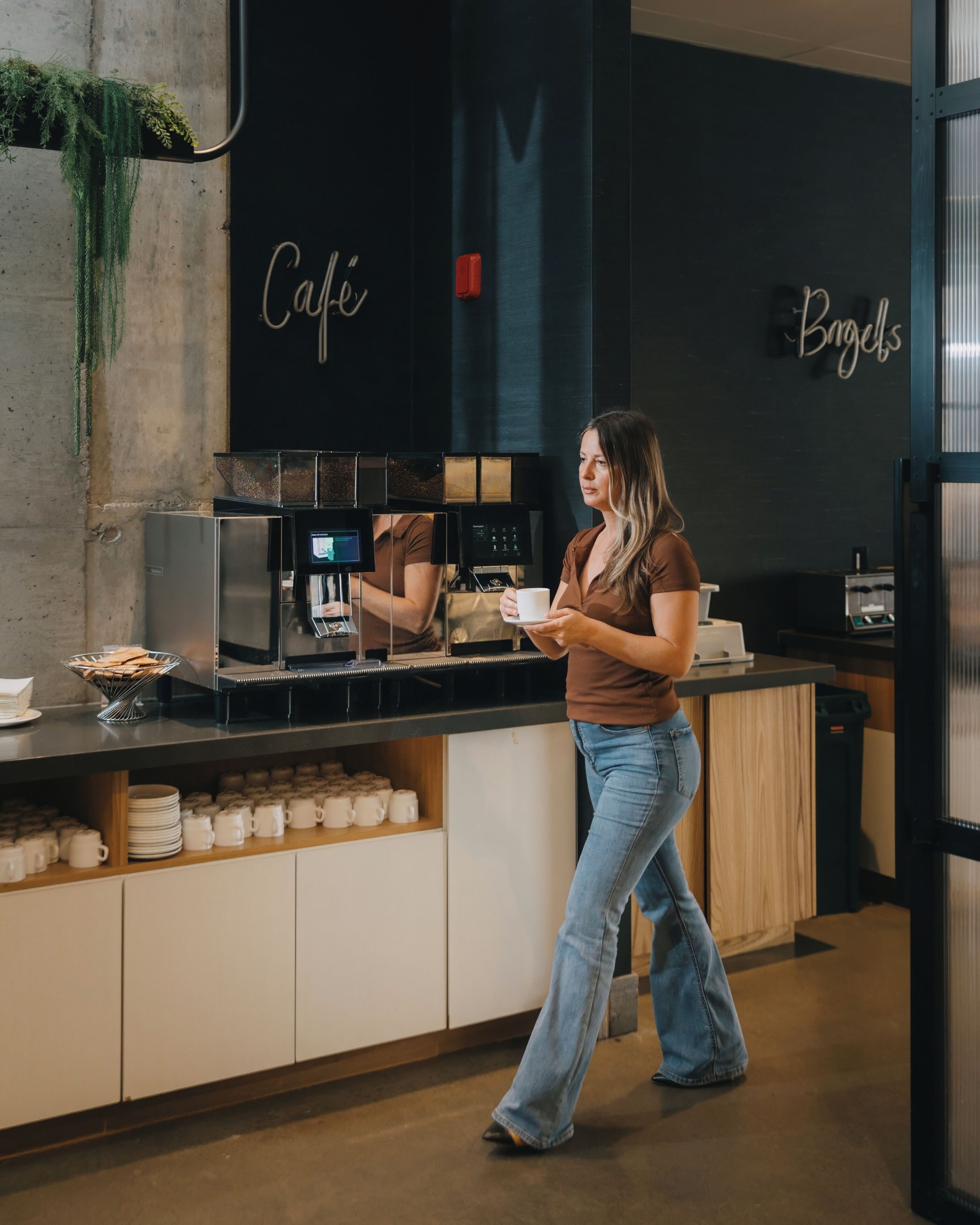 Una mujer con camisa marrón y jeans azules está de pie junto a una estación de café, con una máquina de café y una taza. La estación tiene un mostrador negro y un cartel que dice "Cafe".