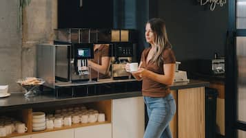 Una mujer con camisa marrón y jeans azules está de pie junto a una estación de café, con una máquina de café y una taza. La estación tiene un mostrador negro y un cartel que dice "Cafe".