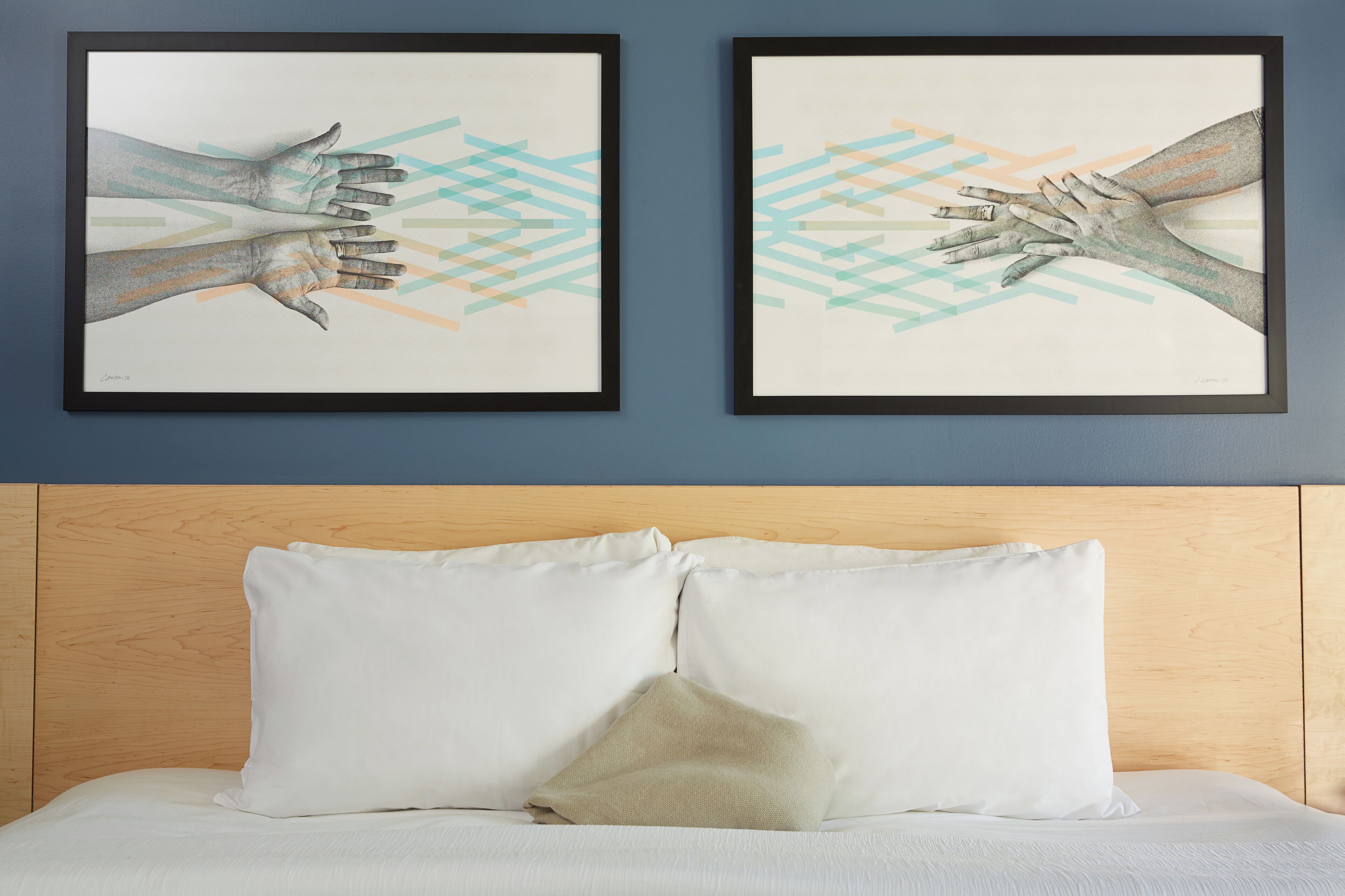 Deux œuvres d’art encadrées représentant des mains tendues, un lit avec une literie blanche et une tête de lit en bois, et un coussin décoratif de couleur beige.