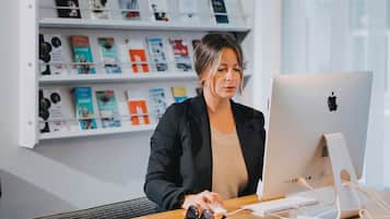 Una mujer sentada en un escritorio con una computadora Apple, en una habitación con estanterías llenas de libros detrás de ella.