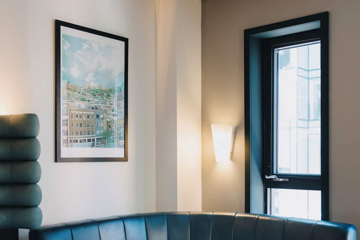Une chambre d’hôtel moderne avec un canapé bleu aux lignes courbes, une table basse ronde, un tableau représentant un paysage urbain encadré, une lampe fixée au mur et une fenêtre donnant sur des immeubles.