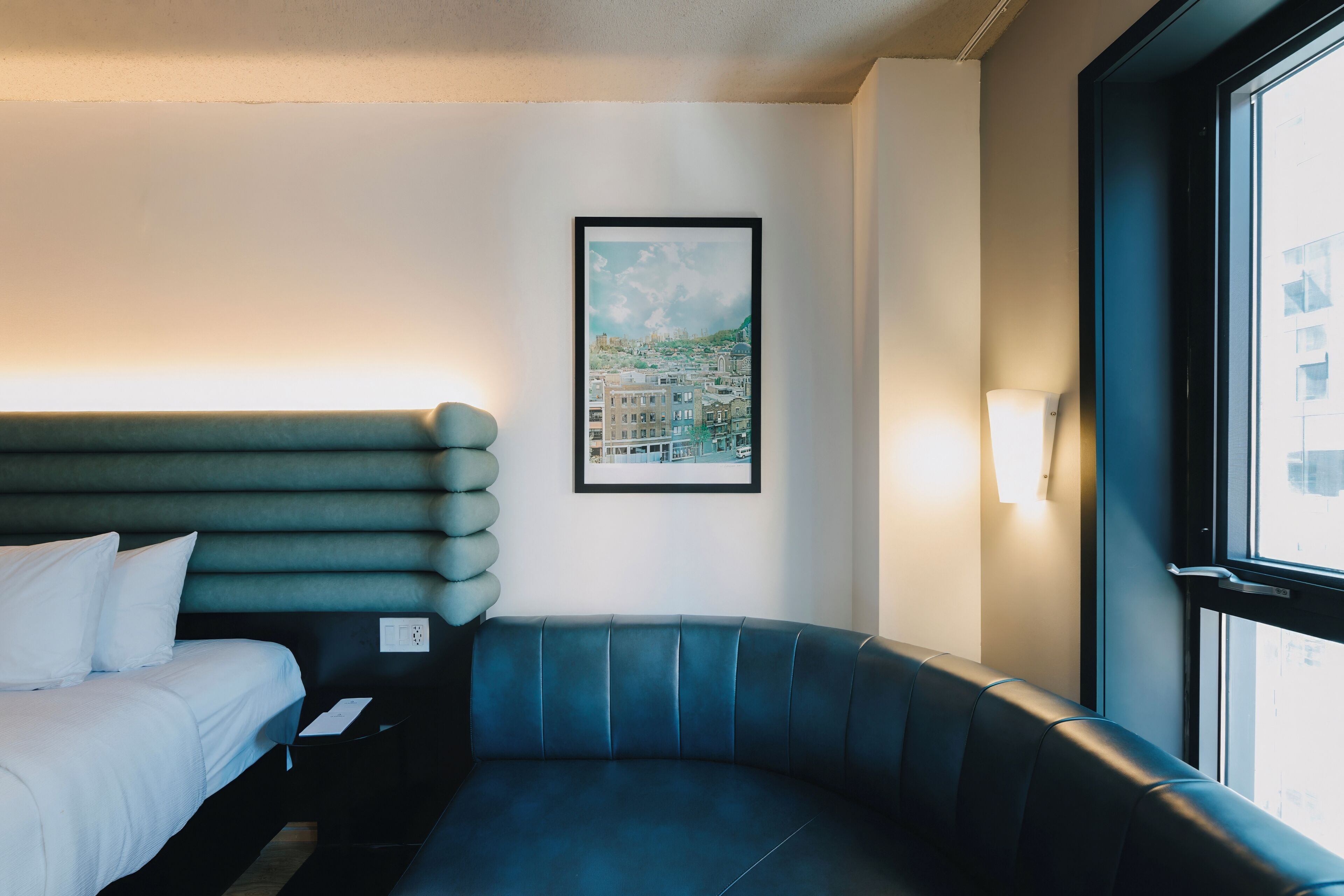 Ein modernes Hotelzimmer mit einem blauen, bogenförmigen Sofa, einem ordentlich bezogenen Bett und einem gerahmten Stadtbild an der Wand.