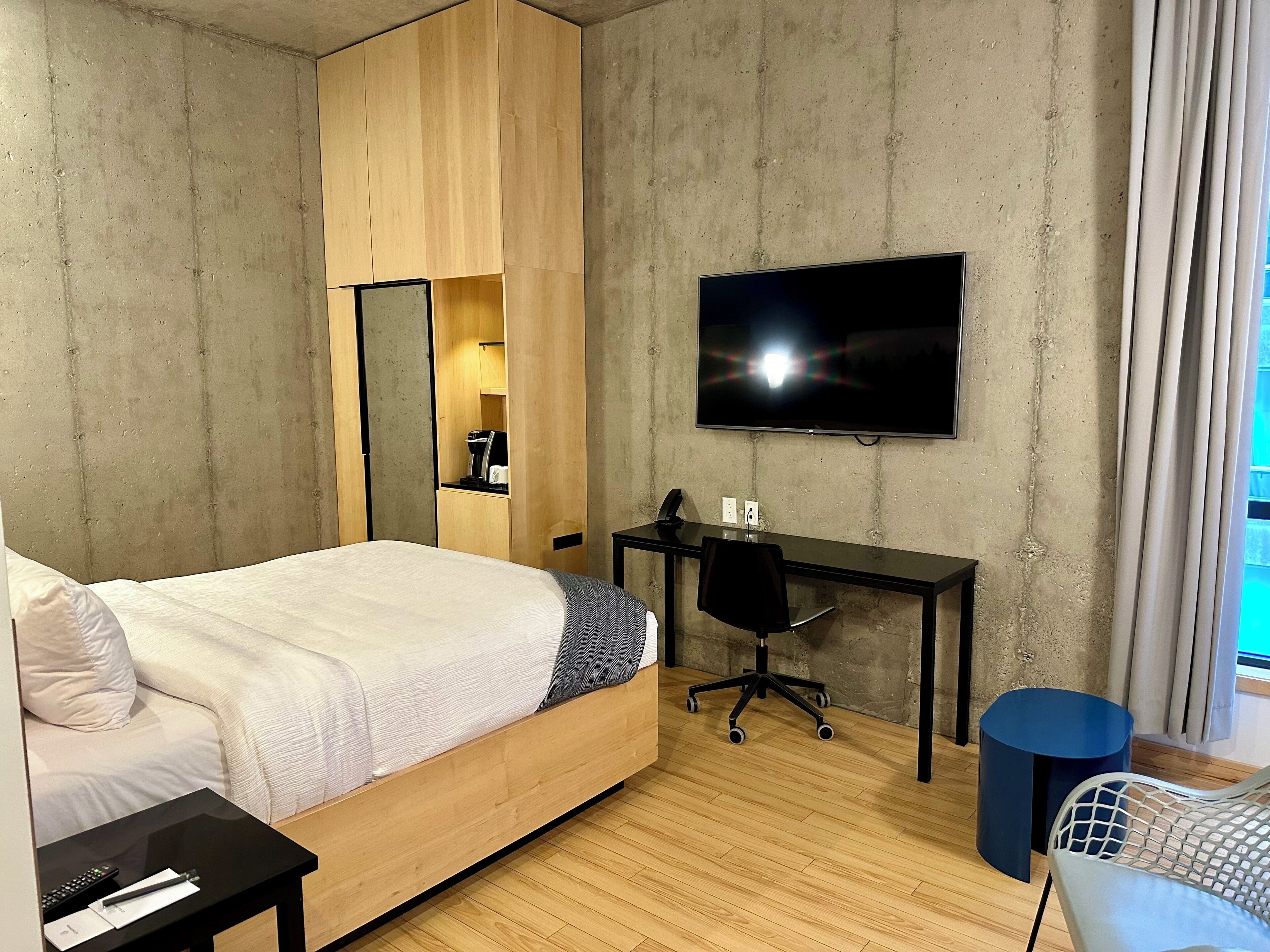 A modern hotel room with a wooden bed, a desk with a chair, a television, and a small blue trash bin.