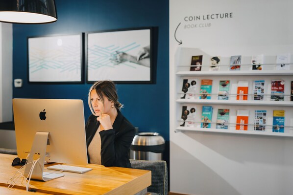 Une femme est assise à un bureau devant un ordinateur Apple iMac, avec un casque et un clavier. Des œuvres d’art encadrées ornent le mur, et une bibliothèque remplie de livres se trouve à proximité.