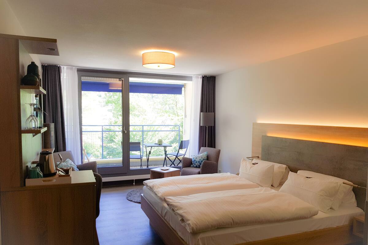 Habitación de hotel con una cama grande, un escritorio, una silla y un balcón con vistas a árboles.