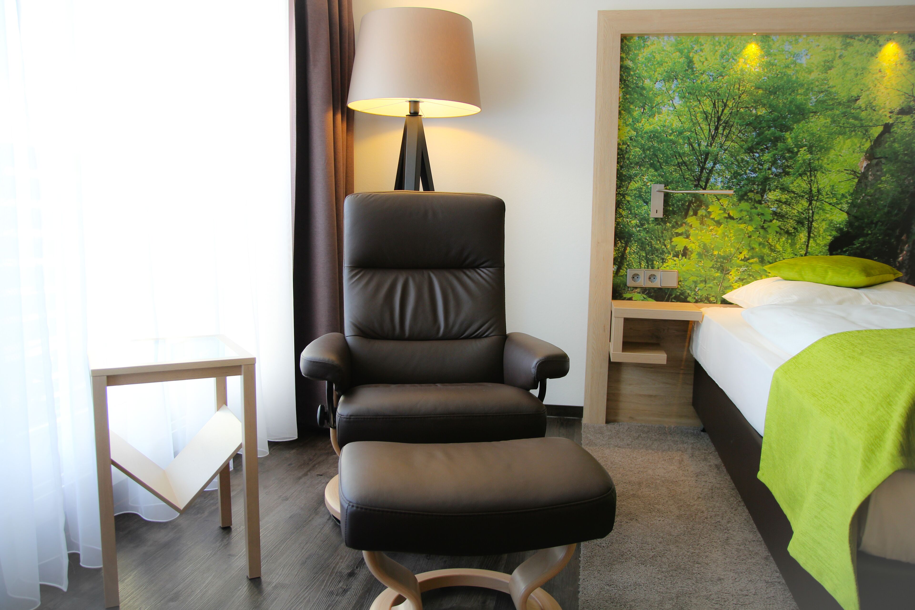 Ein Hotelzimmer mit schwarzem Ledersessel, einem kleinen Tisch, einem Bett mit grüner Decke und einem Wandbild eines Waldes.