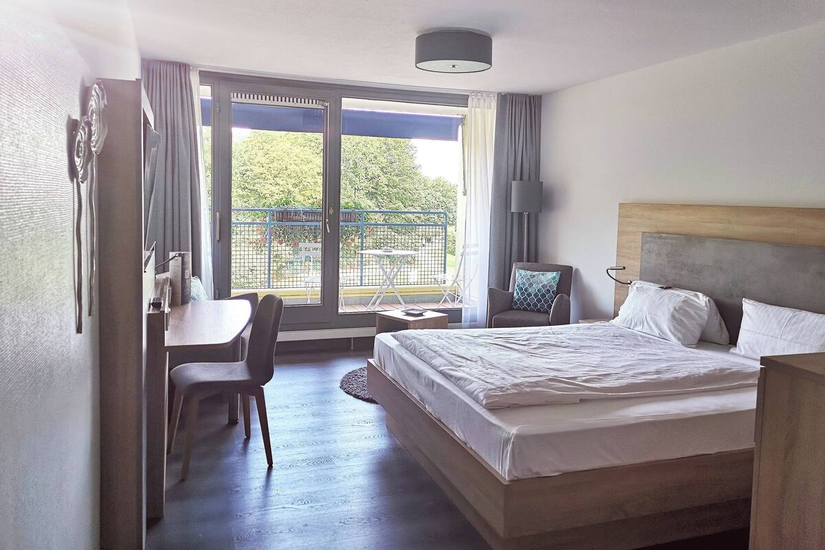 Habitación de hotel con cama, escritorio, silla, una mesita y balcón con vista a la vegetación.