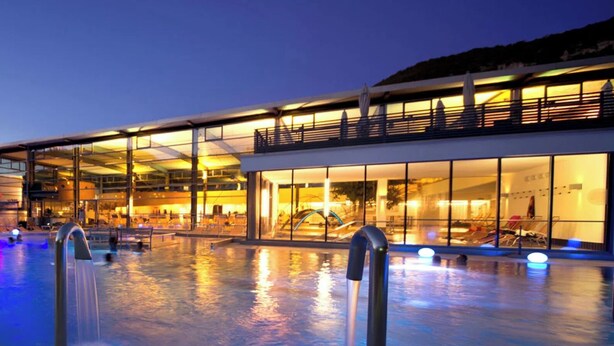 A modern hotel with a large outdoor pool, glass facade, and a well-lit interior.