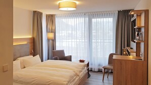 A hotel room with a large bed, a desk, a chair, and a view of the outdoors through sheer curtains.