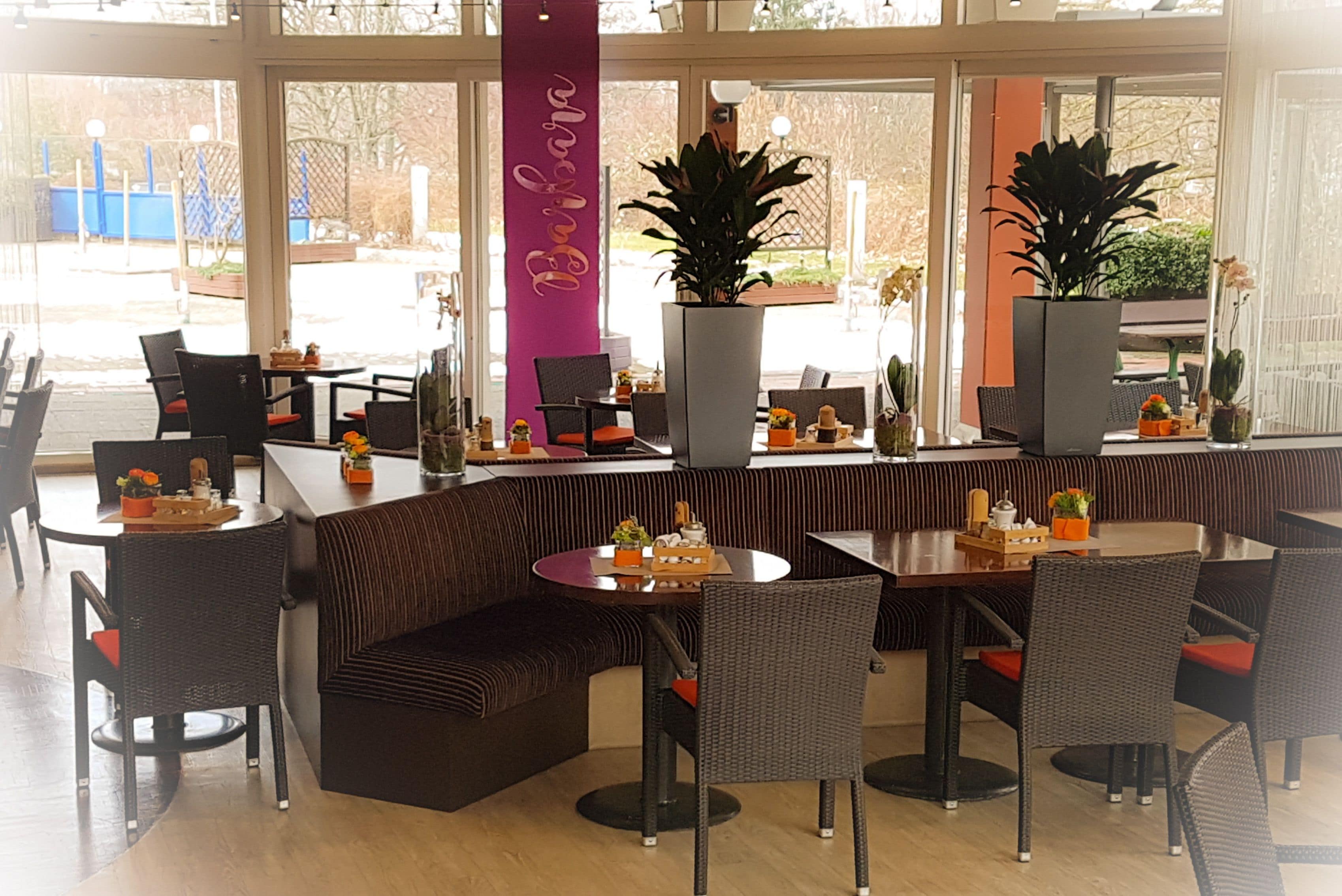 Un restaurante con una decoración moderna, que ofrece una variedad de opciones de asientos, como mesas y cabinas, y grandes ventanales con vistas al exterior.